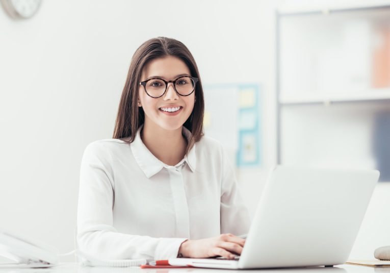 Young woman working at office desk, she is typing on a laptop and smiling, job and career concept